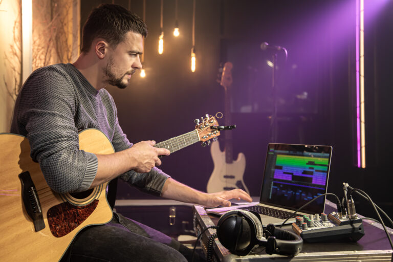 A man with a guitar records sound in a music studio using a special program on his laptop against a blurred dark background.